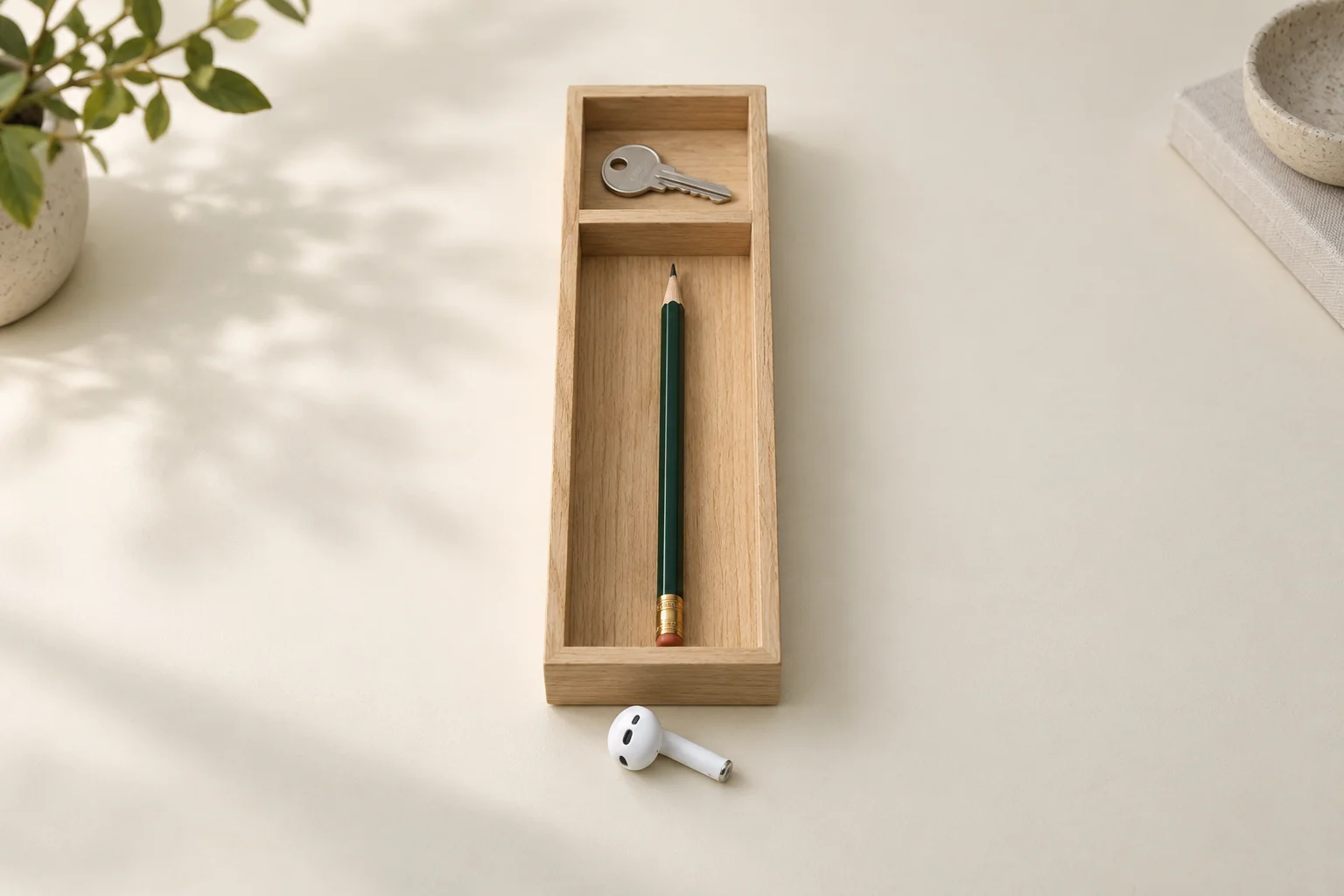 Illustrative divided wooden tray with a key in the smaller compartment, a green pencil in the longer compartment and one white wireless earbud nearby